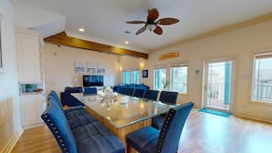 A spacious dining area with a large glass table surrounded by blue upholstered chairs, a ceiling fan, and a view of the outside through large windows.