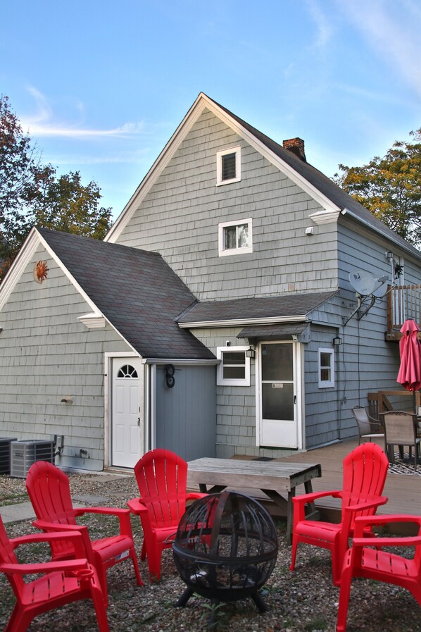 A two-story house with a gray exterior, a front door, and a small porch area with red Adirondack chairs and a fire pit.