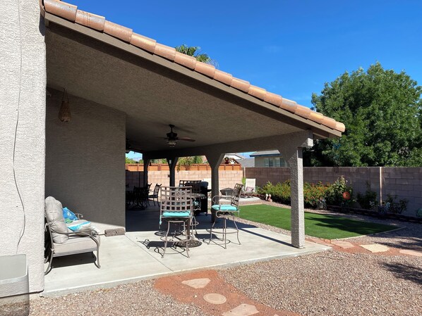 Un patio techado con mobiliario exterior, ventilador de techo y vista al jardín trasero.