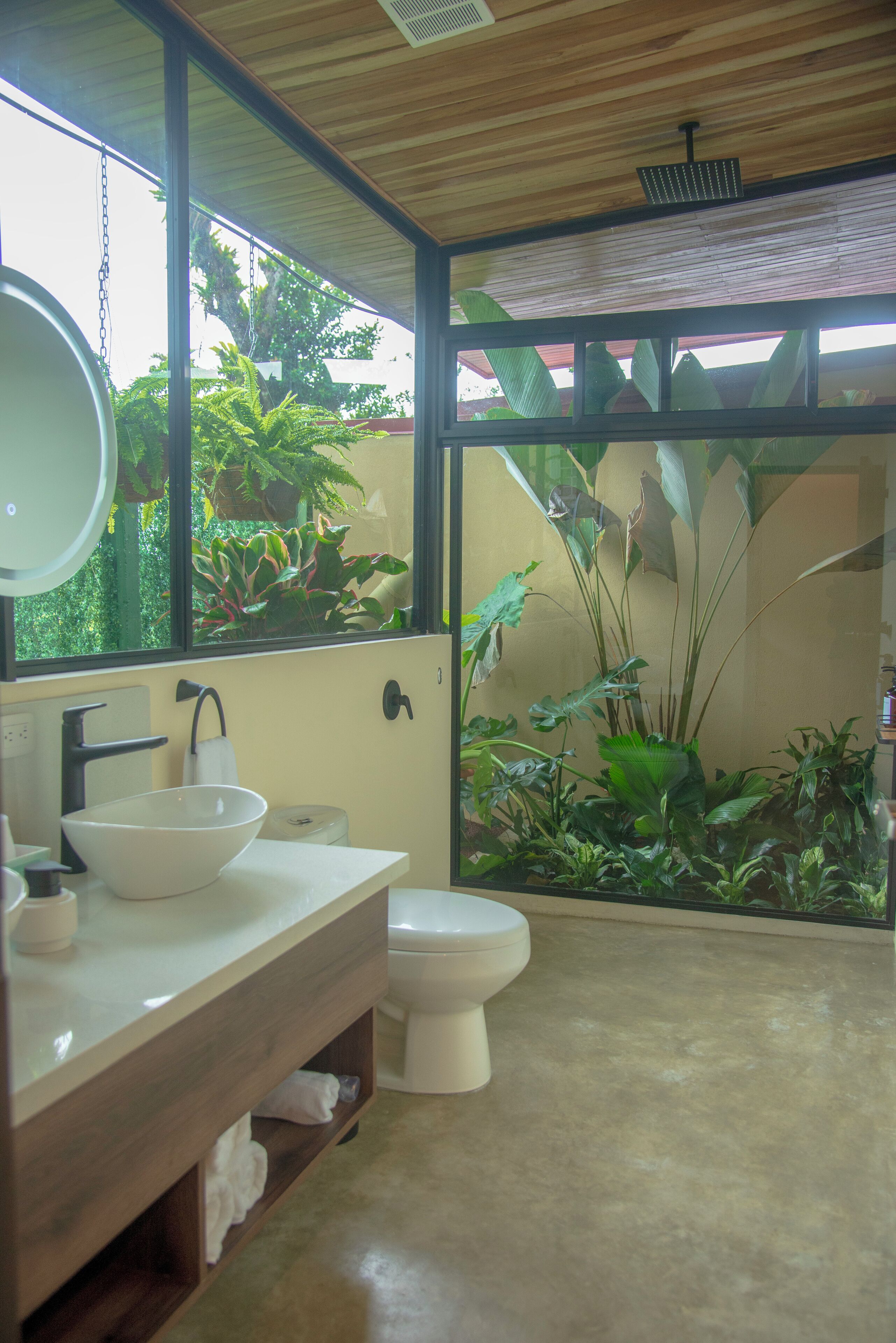 Une salle de bain avec un meuble-lavabo en bois, un lavabo blanc et des toilettes. Une grande fenêtre offre une vue sur la verdure à l’extérieur.