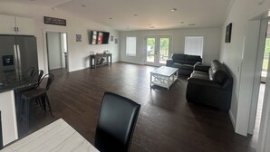 A spacious living room with a dark wood floor, a black leather sofa, a white coffee table, and a flat-screen TV mounted on the wall.