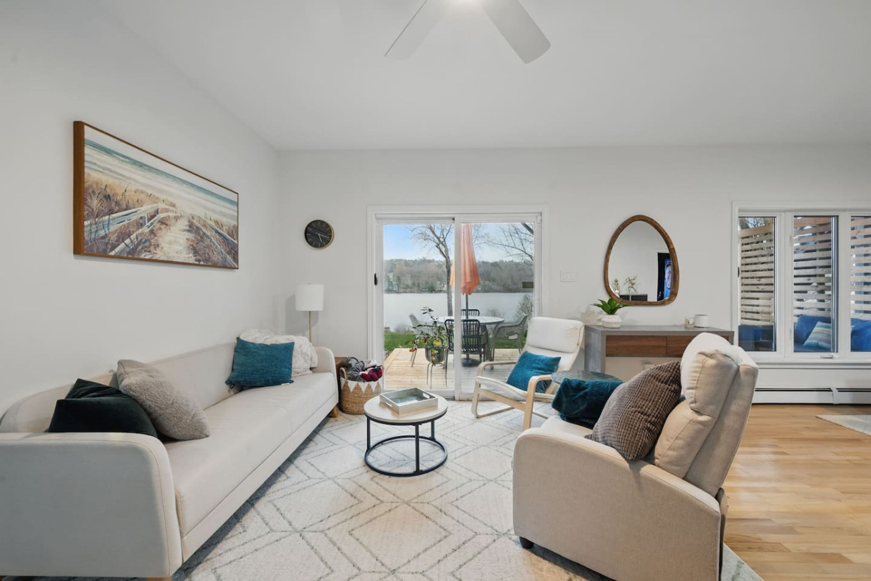 A modern living room with a sofa, armchair, and a coffee table. There is a painting on the wall, a window with blinds, and a view of the outdoors.