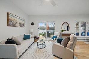 A modern living room with a sofa, armchair, and a coffee table. There is a painting on the wall, a window with blinds, and a view of the outdoors.