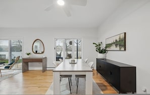 Uma sala de estar moderna com uma mesa de jantar, cadeiras, um sofá, um rack para TV e uma janela grande com vista para o exterior.