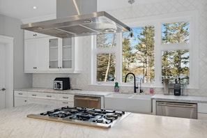 A modern kitchen with a gas stove, white cabinets, a built-in oven, and a window with a view of trees. - Private Lakefront Mansion on Schmidt Lake (Hammonds Plains)