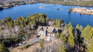A large house with a garage and driveway, surrounded by dense forest and a lake.