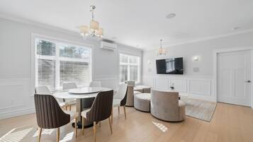 A modern living room with a dining area, large windows with blinds, a flat-screen TV mounted on the wall, and a round coffee table.