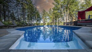 A rectangular swimming pool with clear blue water, surrounded by a stone deck and wooden lounge chairs.