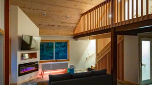 A modern living room with a wooden ceiling, a fireplace, a large window, and a staircase.