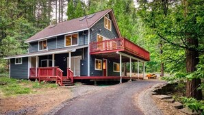 A two-story cabin with a red porch and a gravel driveway, surrounded by forest.