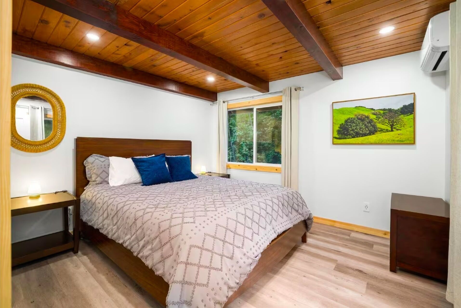 A bedroom with a wooden ceiling, a bed with patterned bedding, a bedside table, a window with a view of greenery, and a landscape painting on the wall.
