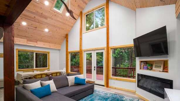 A spacious living room with a gray sectional sofa, a blue rug, a fireplace, and large windows offering views of the outdoors.