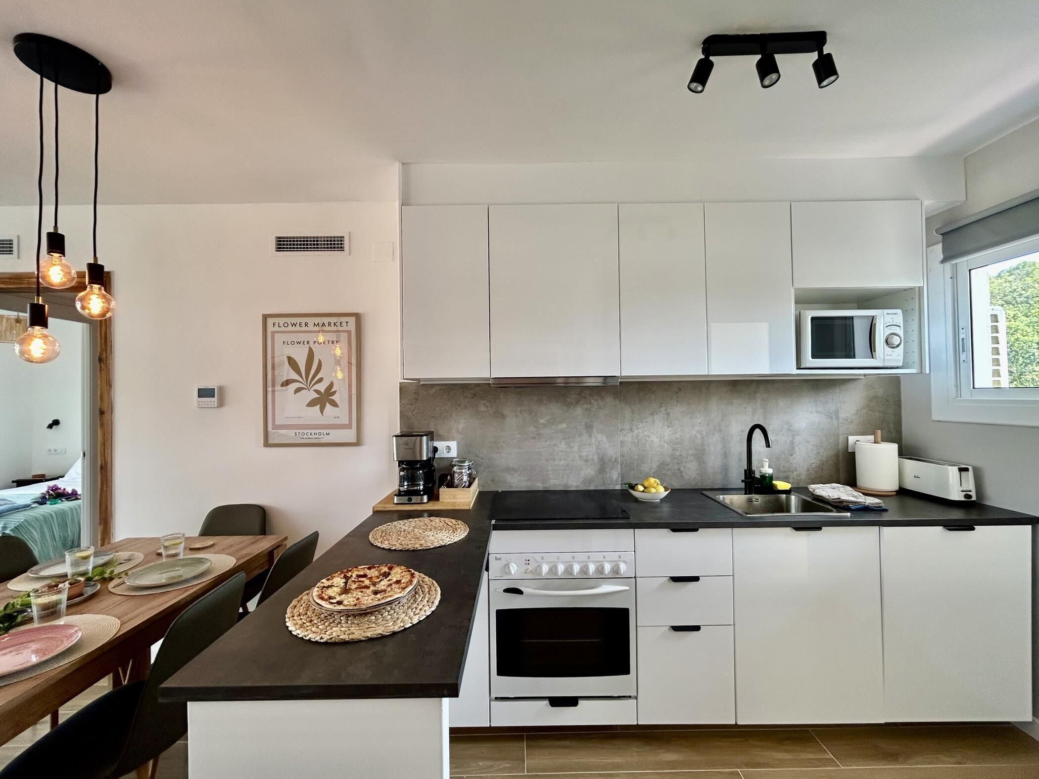 Una cocina moderna con armarios blancos, encimera negra y horno empotrado. Hay un microondas sobre la encimera, un lavabo con grifo y una ventana que proporciona luz natural.