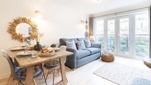 A modern living room with a wooden dining table, grey sofa, and large windows. - 26 Waves - sleeps 4 guests  in 1 bedroom (Watergate Bay)