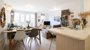 A bright, open-plan living and dining area with a large window, wooden furniture, and a variety of decorative items. - 26 Waves - sleeps 4 guests  in 1 bedroom (Watergate Bay)