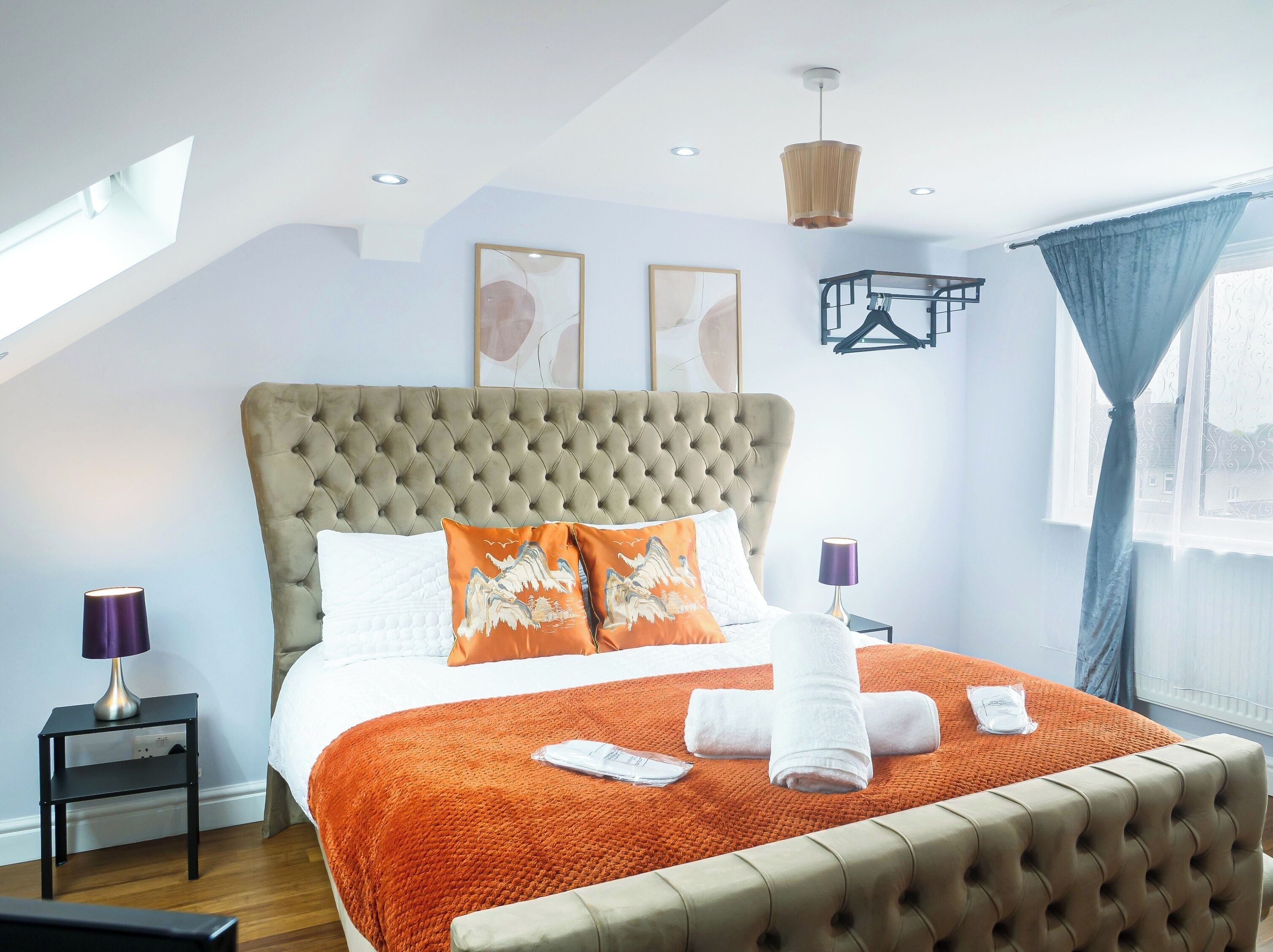 Un dormitorio con cabecera capitonada, una cama grande con manta naranja, dos almohadas, una mesita de noche con lámpara y una ventana con cortinas.