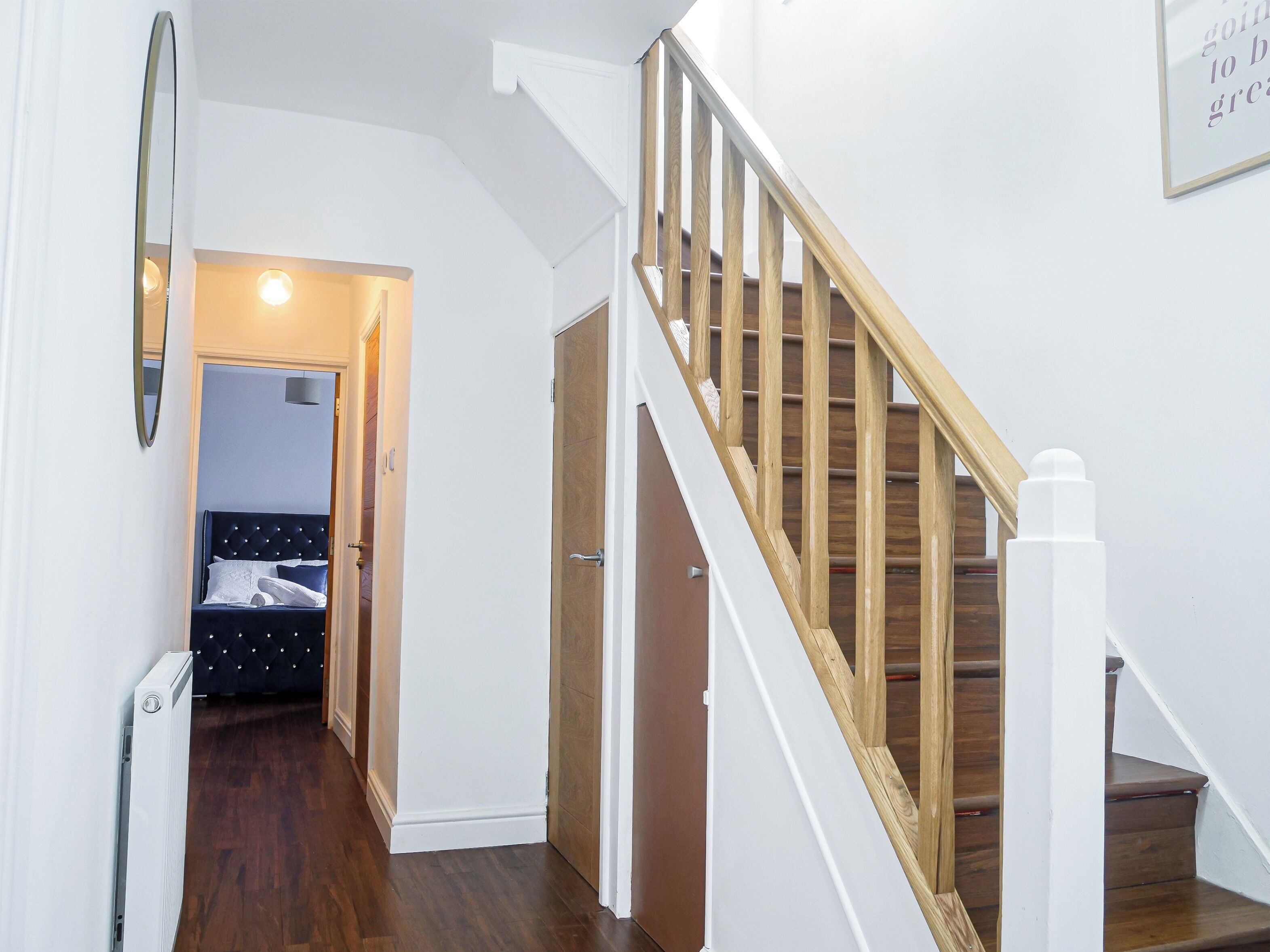 Un escalier avec des rampes en bois et une main courante blanche, menant à une chambre à coucher équipée d’un lit et d’une table de chevet.