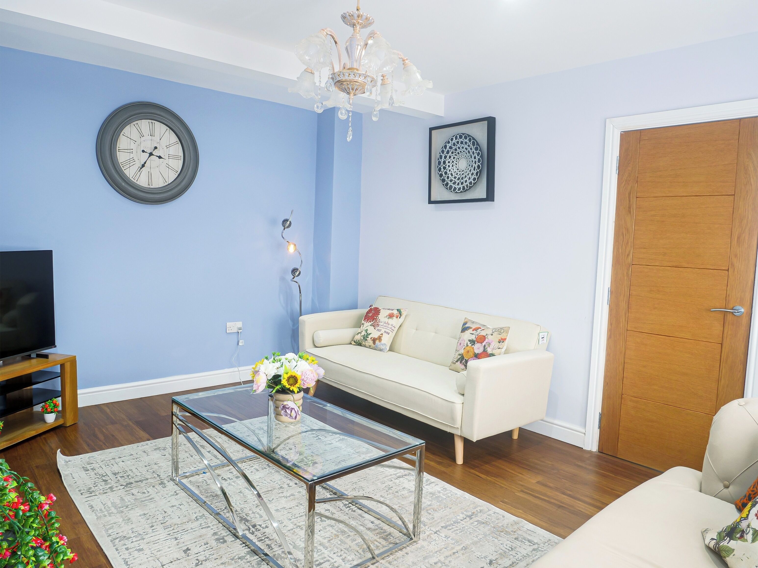 Una sala de estar con un sofá blanco, una mesa de centro de vidrio con flores, un reloj en la pared y una araña.