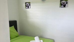 Een kleine, schone kamer met een bed, een groene dekbedovertrek, witte kussens en een handdoek. Er zijn twee wandmontage airconditioners en twee kleine kunstwerken aan de muur.