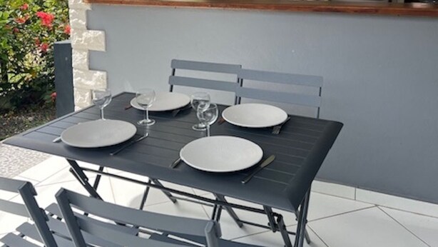 Een terras met eettafel voor vier personen, een keuken zichtbaar door een open deur, en uitzicht op een tuin met roze bloemen.