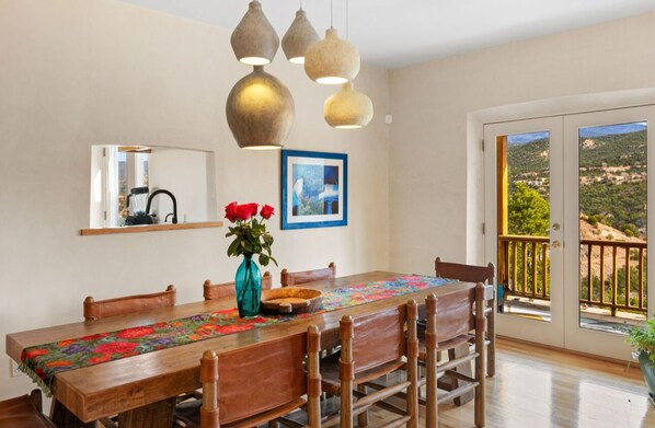 A dining area with a wooden table, chairs, a vase with red flowers, pendant lights, a framed picture, and a view of a mountainous landscape through a glass door. - Pulitzer Hillside Retreat - Hot Tub - Valley Views - Near Plaza & Ski Santa Fe (Santa Fe)