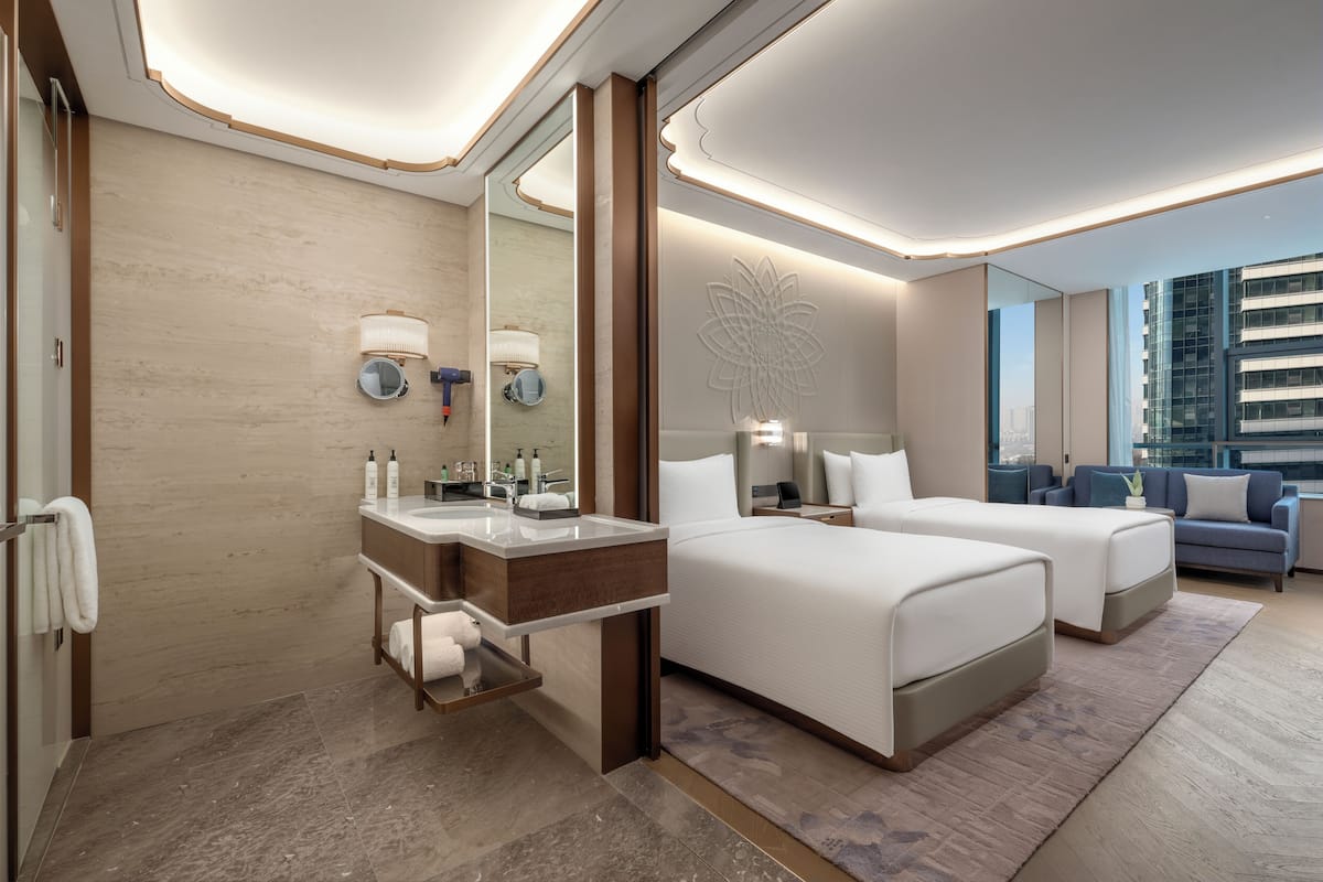 A modern hotel room with a large bed, a separate sofa, a bathroom with a vanity, and a city view.