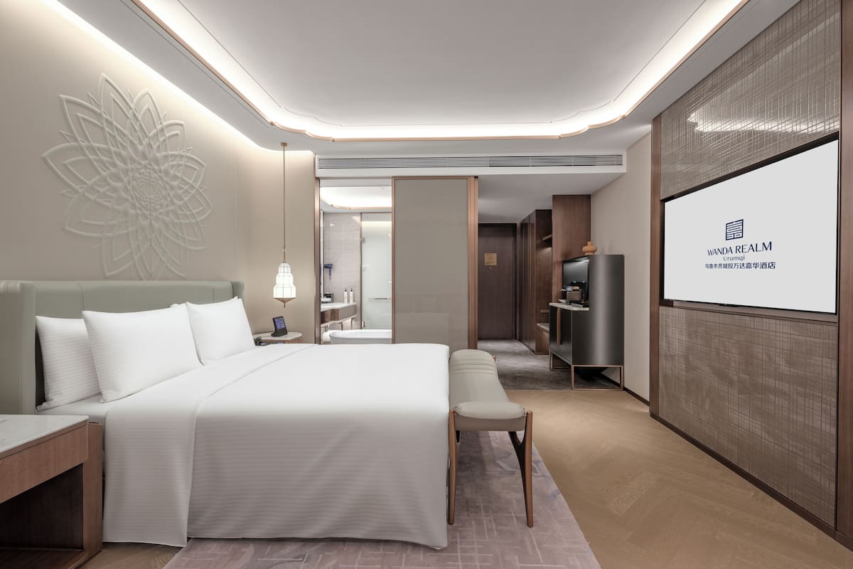 A modern hotel room with a large bed, bedside tables, a desk, a bench, a TV, and a decorative wall piece.