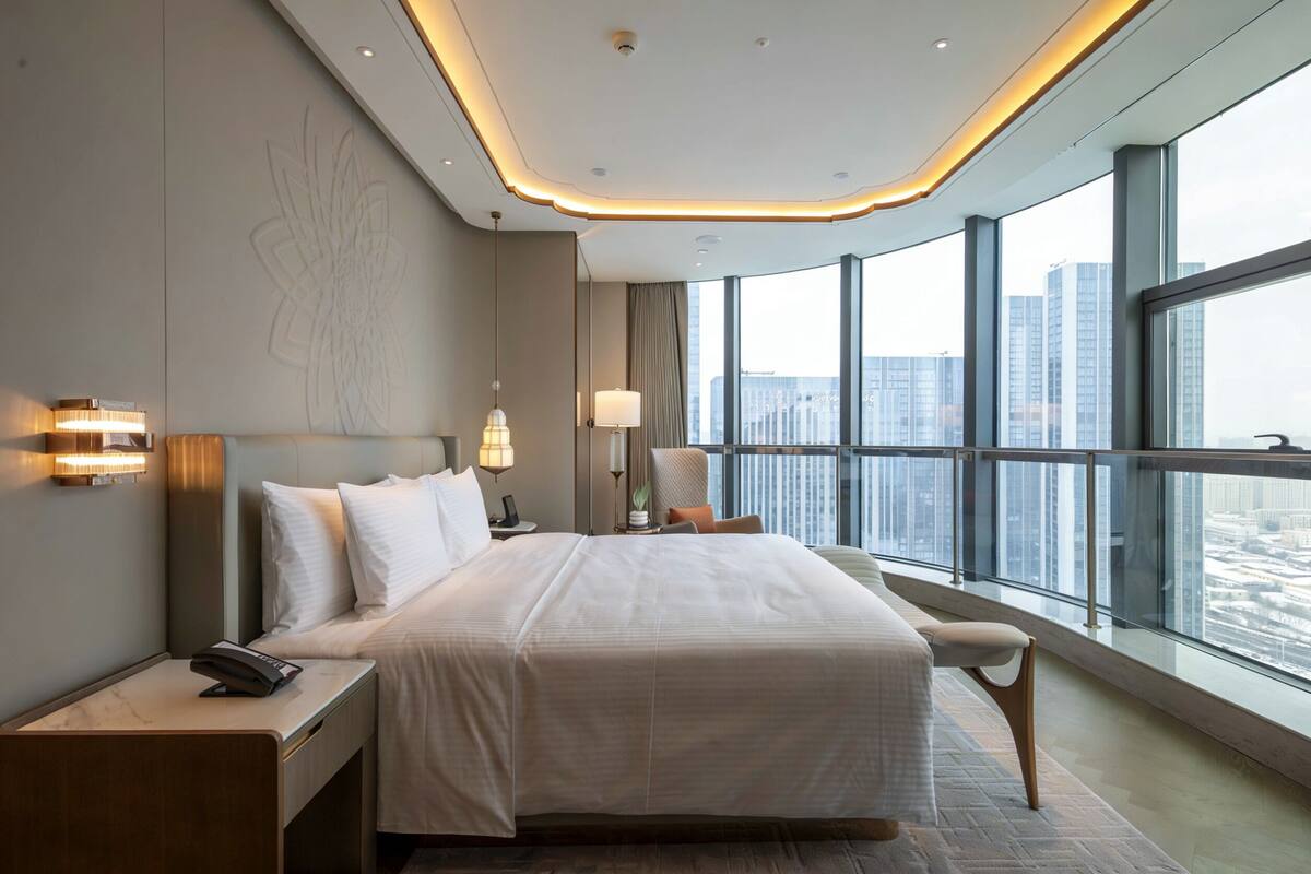 A modern hotel room with a large bed, bedside tables, a nightstand, a chair, and a view of the city through large windows.