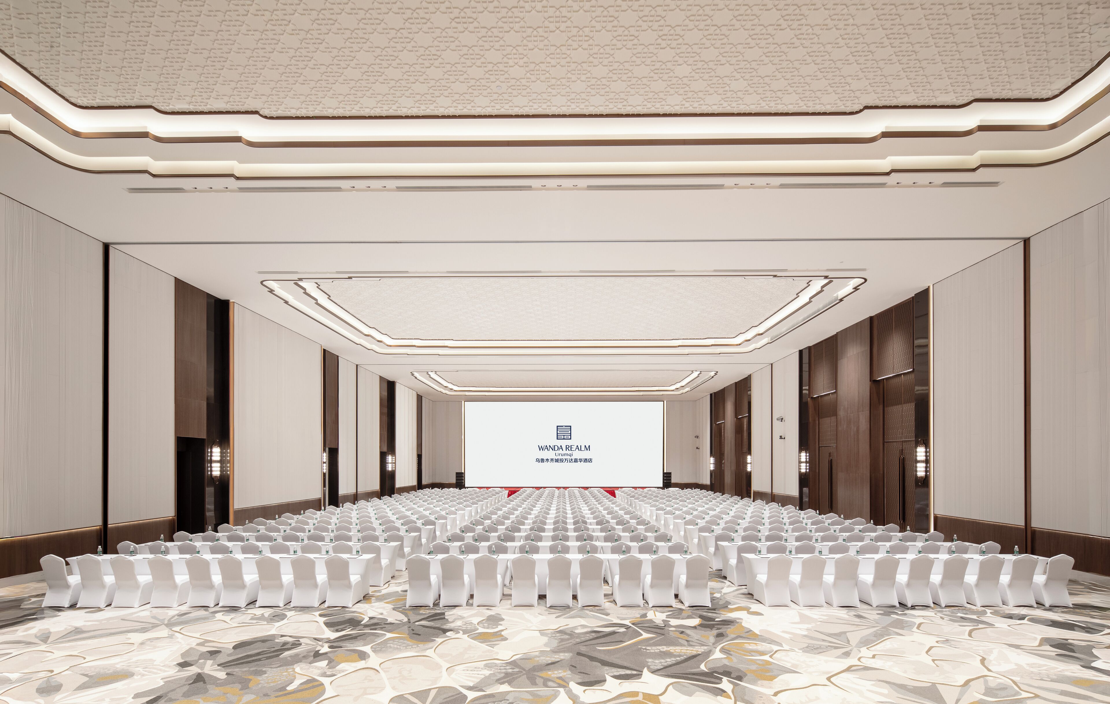 Une salle de conférence avec des rangées de chaises blanches, un grand écran à l’avant et un tapis à motifs.