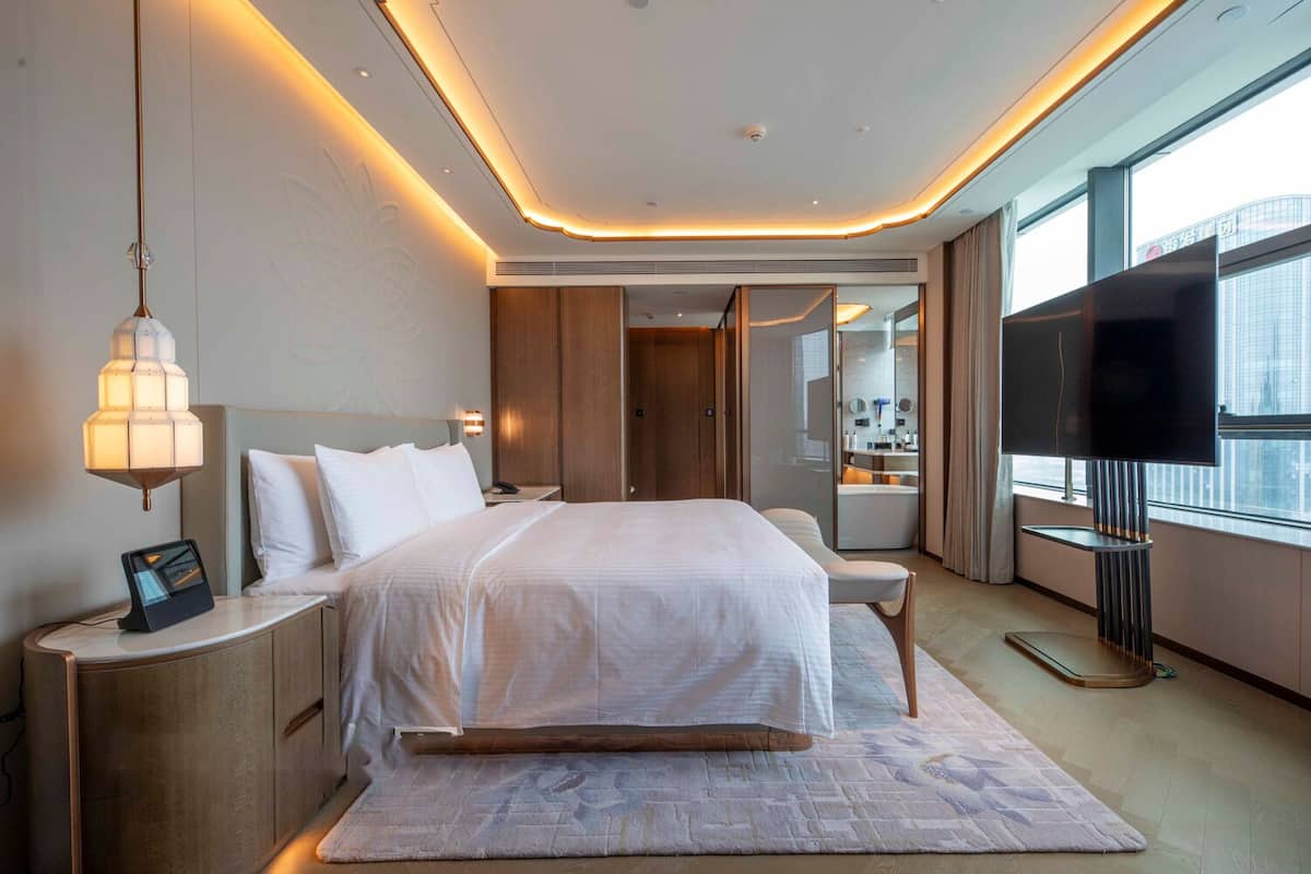 A modern hotel room with a large bed, bedside tables, a flat-screen TV, and a view of the city through large windows.