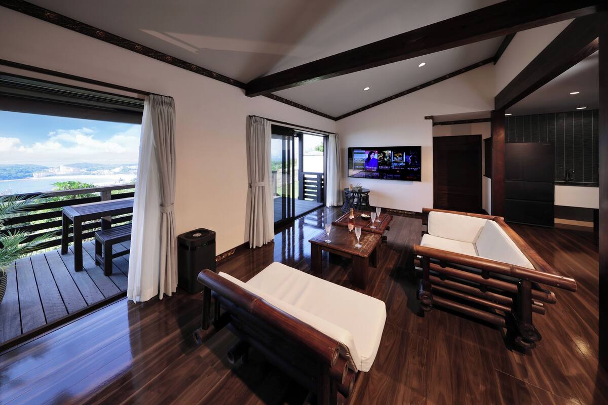 A modern living room with a wooden floor, a sofa, a bench, a TV, and a balcony with a view of the city.
