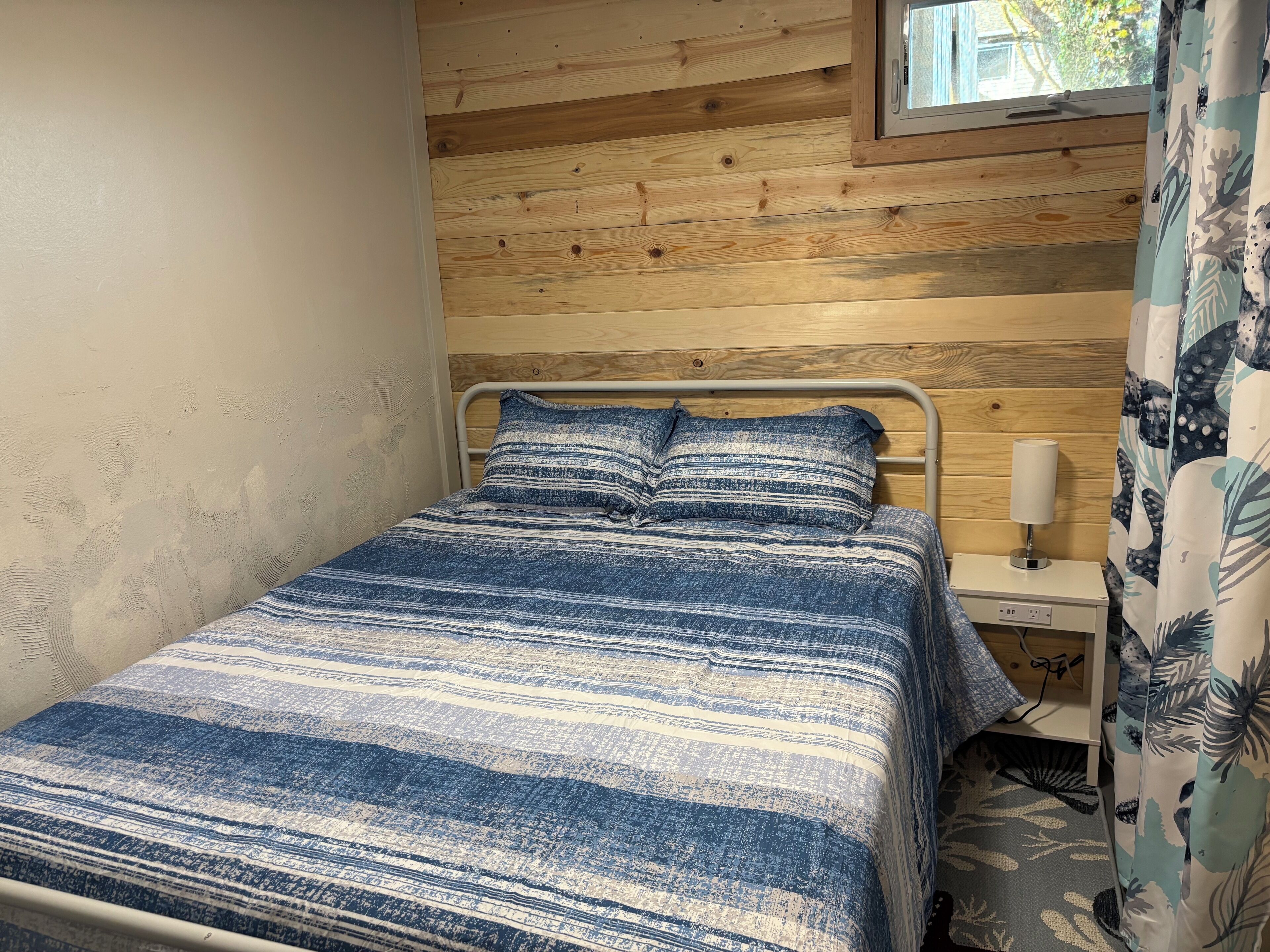 A small, cozy room with a bed, a nightstand, and a window with wooden walls.