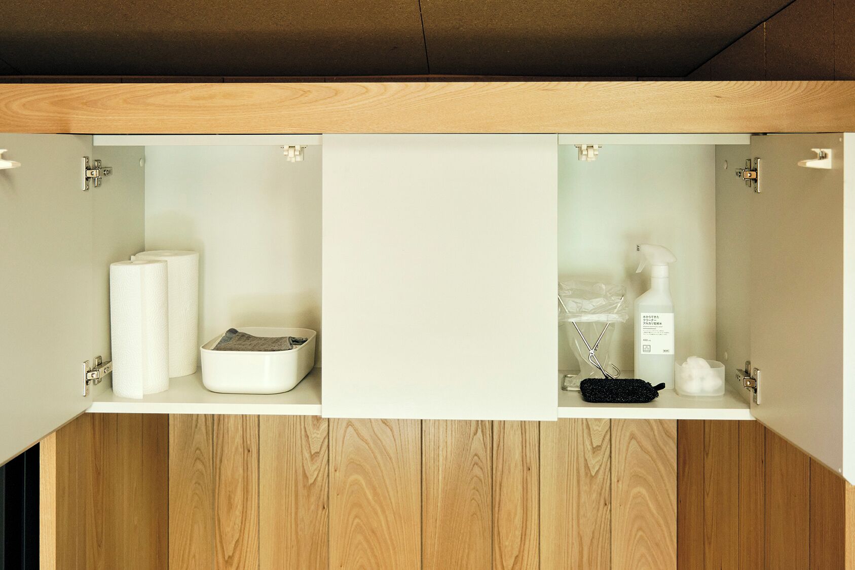A wooden cabinet with two compartments; one holds toilet paper and a tissue box, the other contains a black brush and a white bottle.