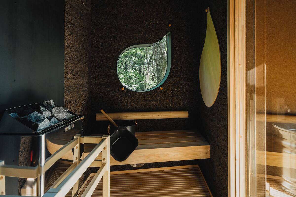 Un sauna avec un banc en bois, un grand récipient noir et une fenêtre donnant sur les arbres.