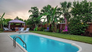 Una piscina con área techada, sillas de descanso y plantas tropicales.