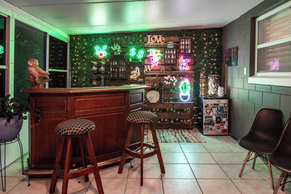 A bar area with stools, a wooden counter, and a wall decorated with neon signs and stickers.