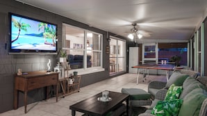 Una sala de estar con televisor, una mesa de comedor, muebles de mimbre y vistas a través de un ventanal corredizo.