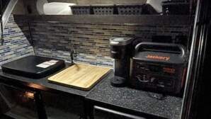Une petite cuisine équipée avec un plan de travail noir, une station d’alimentation portable Jackery, une planche à découper et un évier.
