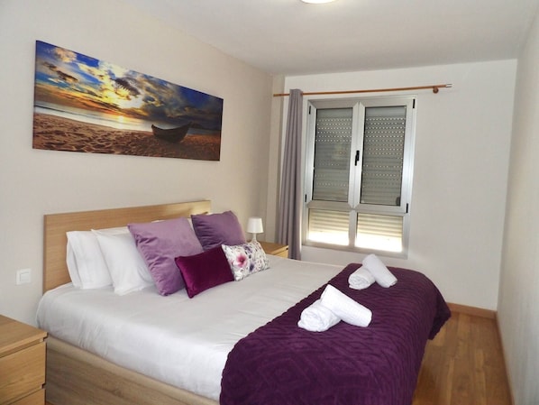 Een slaapkamer met een bed, kussens, een deken, een schilderij, een raam en een gordijn.