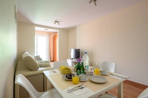 Un comedor con una mesa dispuesta para dos personas, incluyendo platos, cubiertos y bebidas. Se ve un sofá y un televisor al fondo.