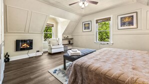 A bedroom with a large bed, a ceiling fan, a fireplace, a TV, and a window with a view of trees.