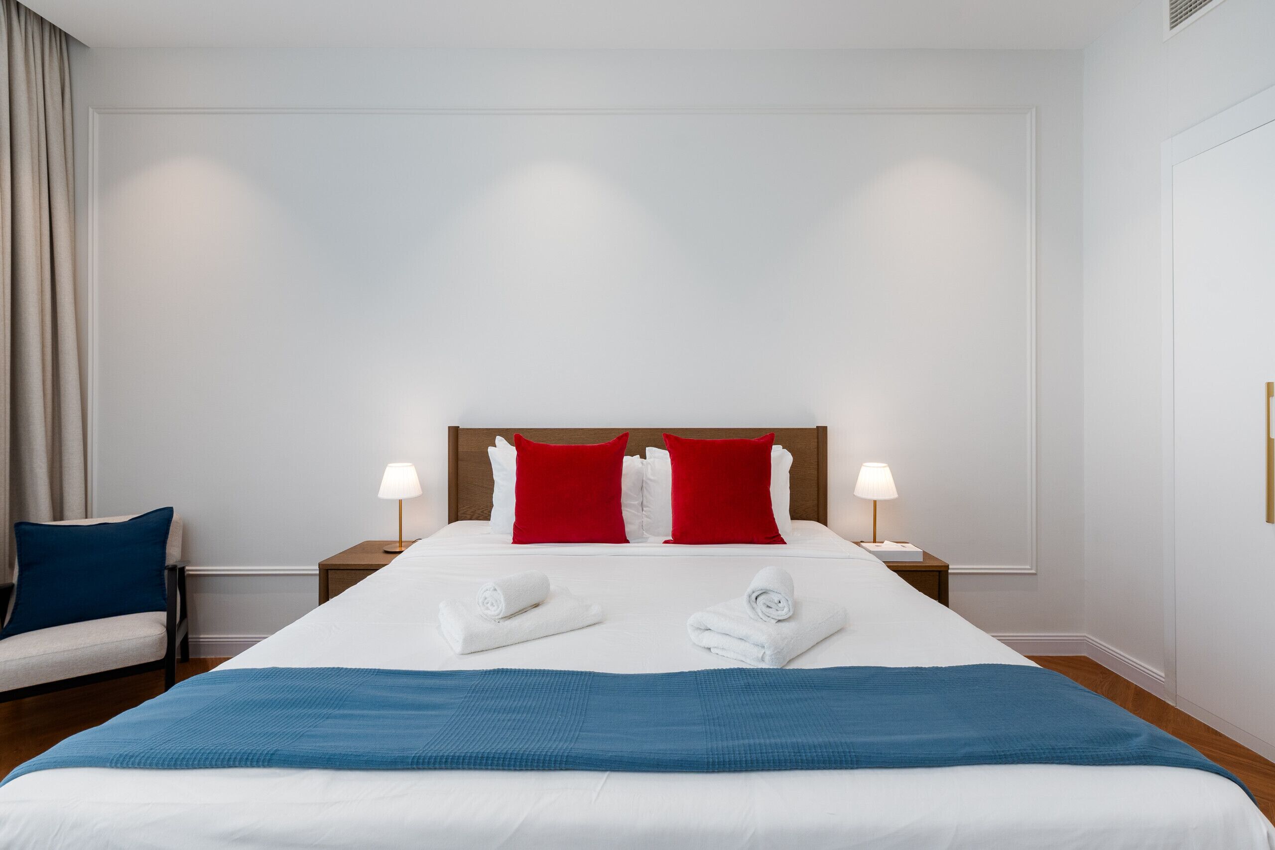 Une chambre d’hôtel avec un grand lit, deux oreillers rouges, du linge de lit blanc, deux lampes de chevet et un plaid bleu.
