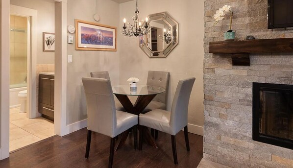 A dining area with a round glass table, two grey upholstered chairs, a chandelier, a framed picture, and a stone fireplace.