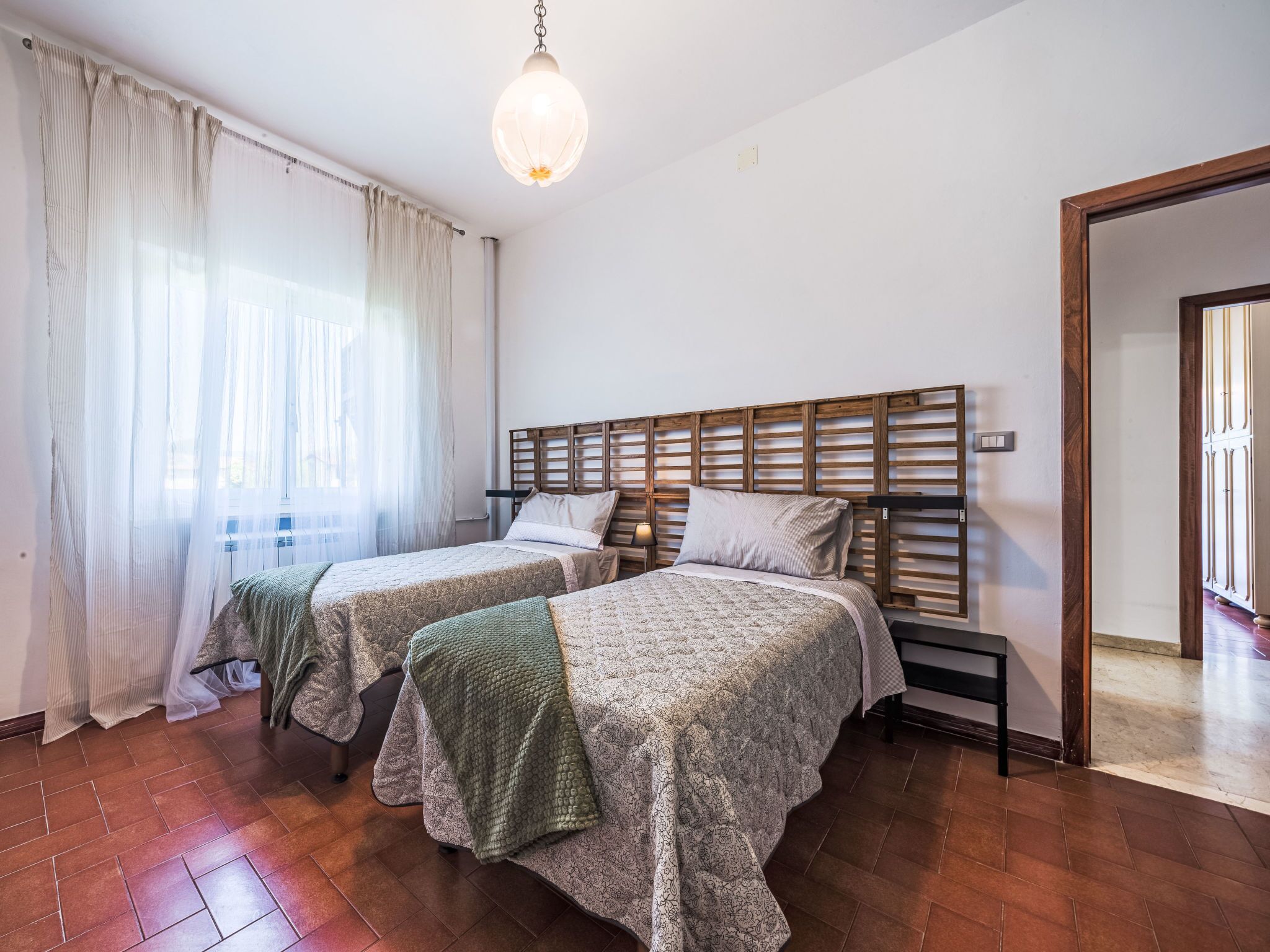 Une chambre avec deux lits, une tête de lit en bois, un luminaire suspendu et une vue sur un couloir.