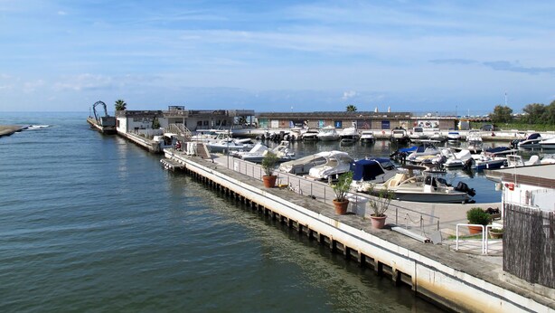 Uma marina com barcos atracados ao longo de um cais, um edifício com letreiros e um céu claro.