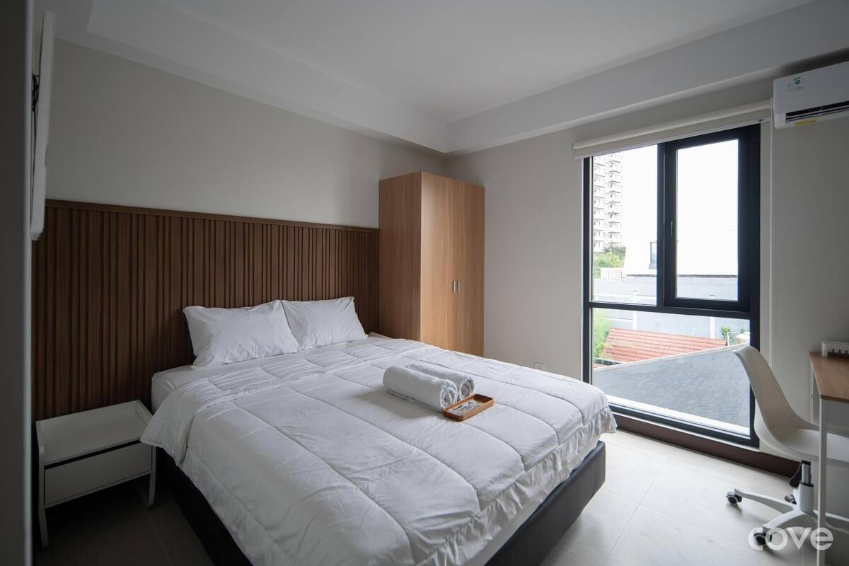 A modern hotel room with a large bed, a wooden wardrobe, and a desk with a chair.