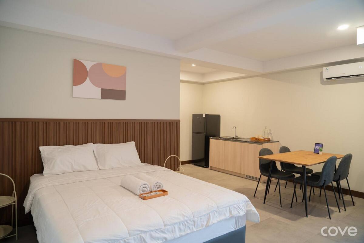 A hotel room with a bed, a small kitchenette, a dining table with chairs, and a wall-mounted air conditioner.