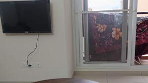 A round wooden table with a green upholstered ottoman, a flat-screen TV mounted on the wall, and a sliding glass door with a floral view.