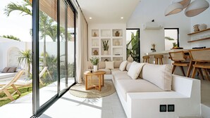 A modern living room with a large window, a white sofa, a dining area with wooden chairs, and a wall with shelves displaying decorative items.