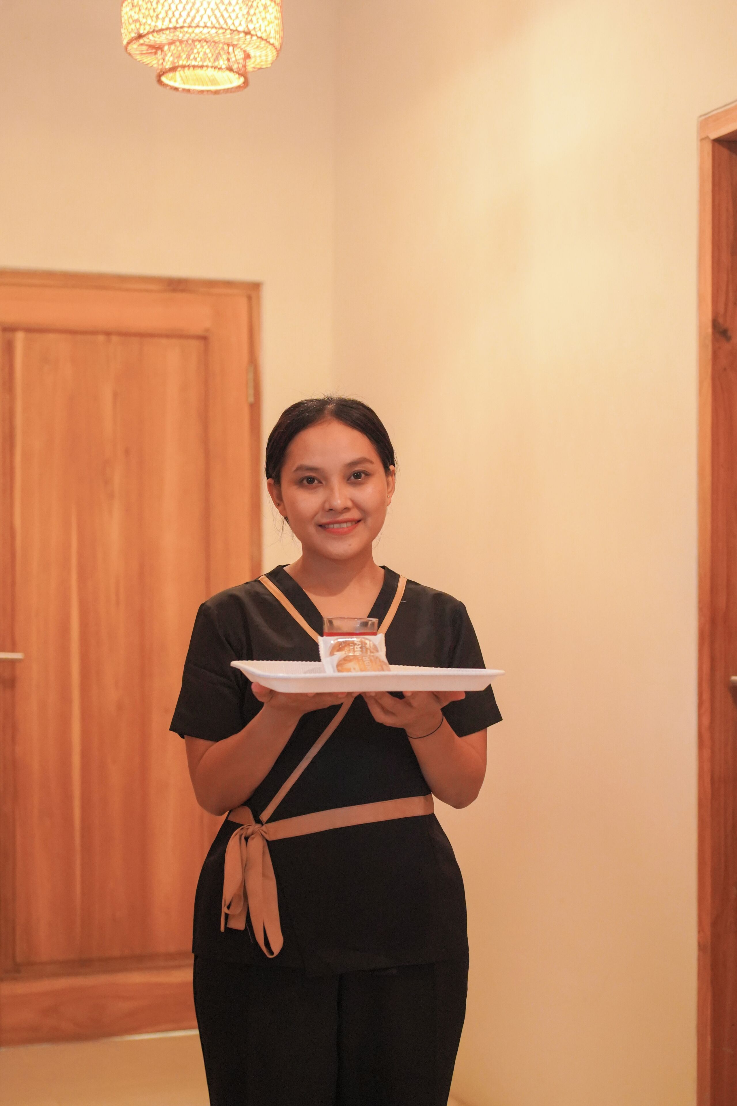 A person holding a tray with a plate and a glass, standing in a hallway with wooden doors and a hanging light fixture.